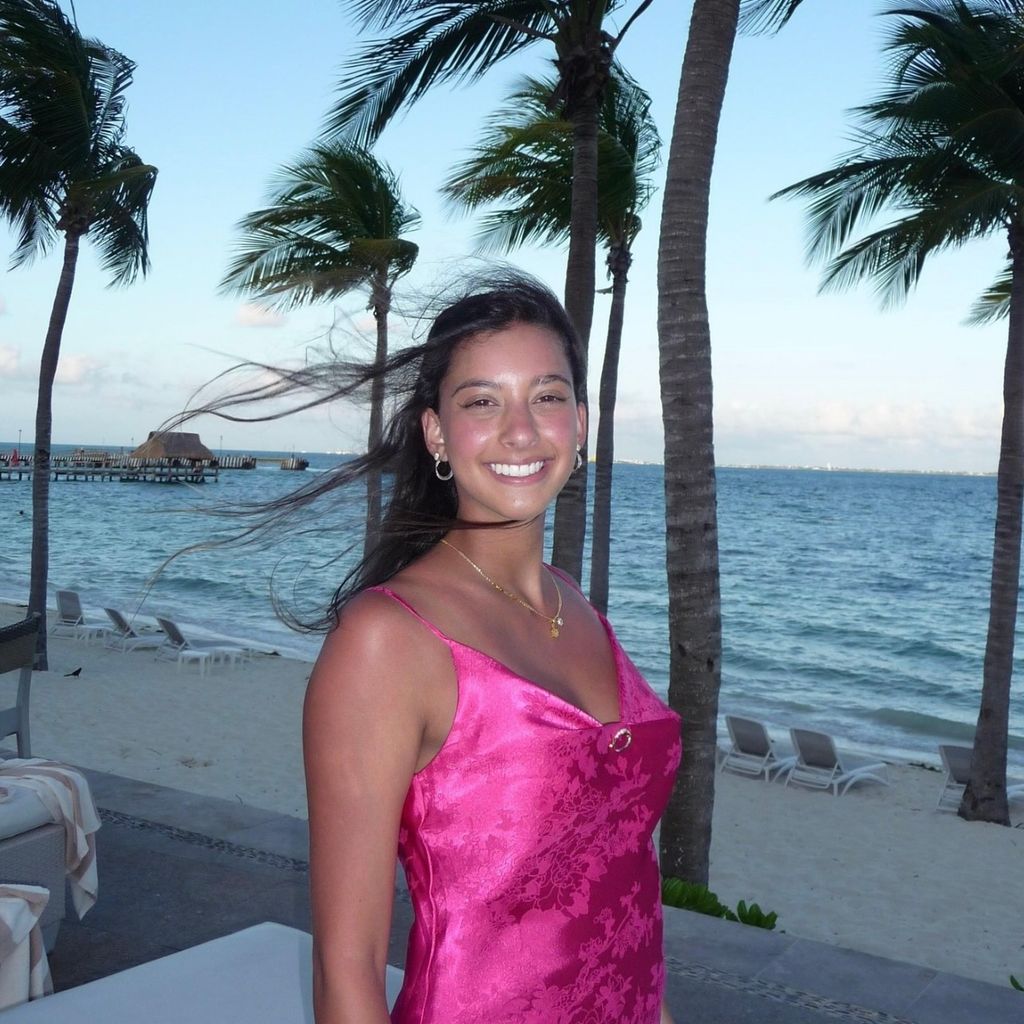 Photo: A person stands on a beachside path at sunset, wearing a hot pink satin dress with thin straps. Tall palm trees, white lounge chairs, and a calm ocean are in the background.