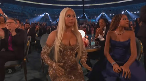 Meme: People seated at a round table in a large, crowded venue with bright stage lights. One person, Beyonce, is wearing a sparkly gold dress and long gloves, another is in a dark blue dress.
