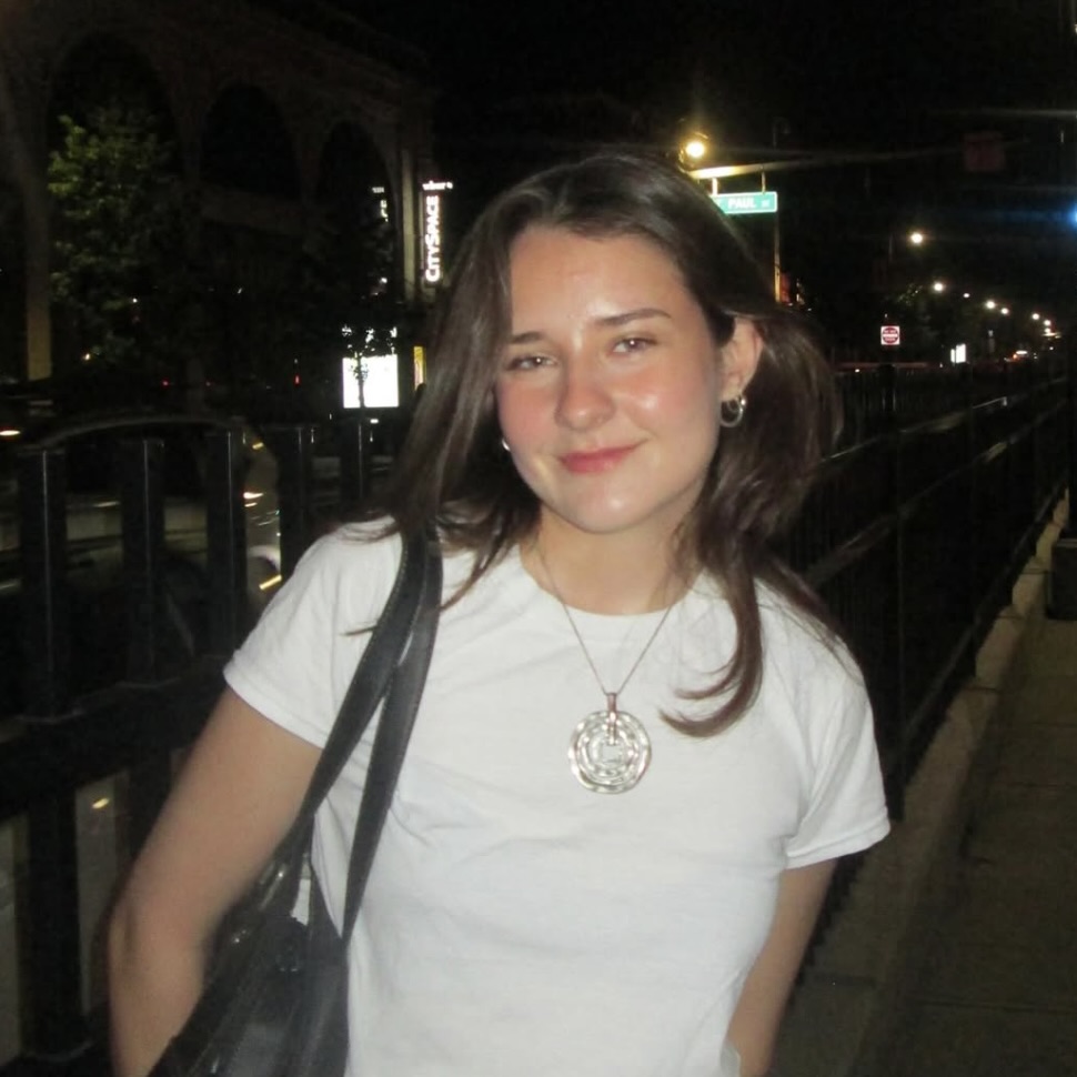 Photo: A person in a white t-shirt and necklace stands outdoors at night on a city sidewalk, carrying a black bag over one shoulder.