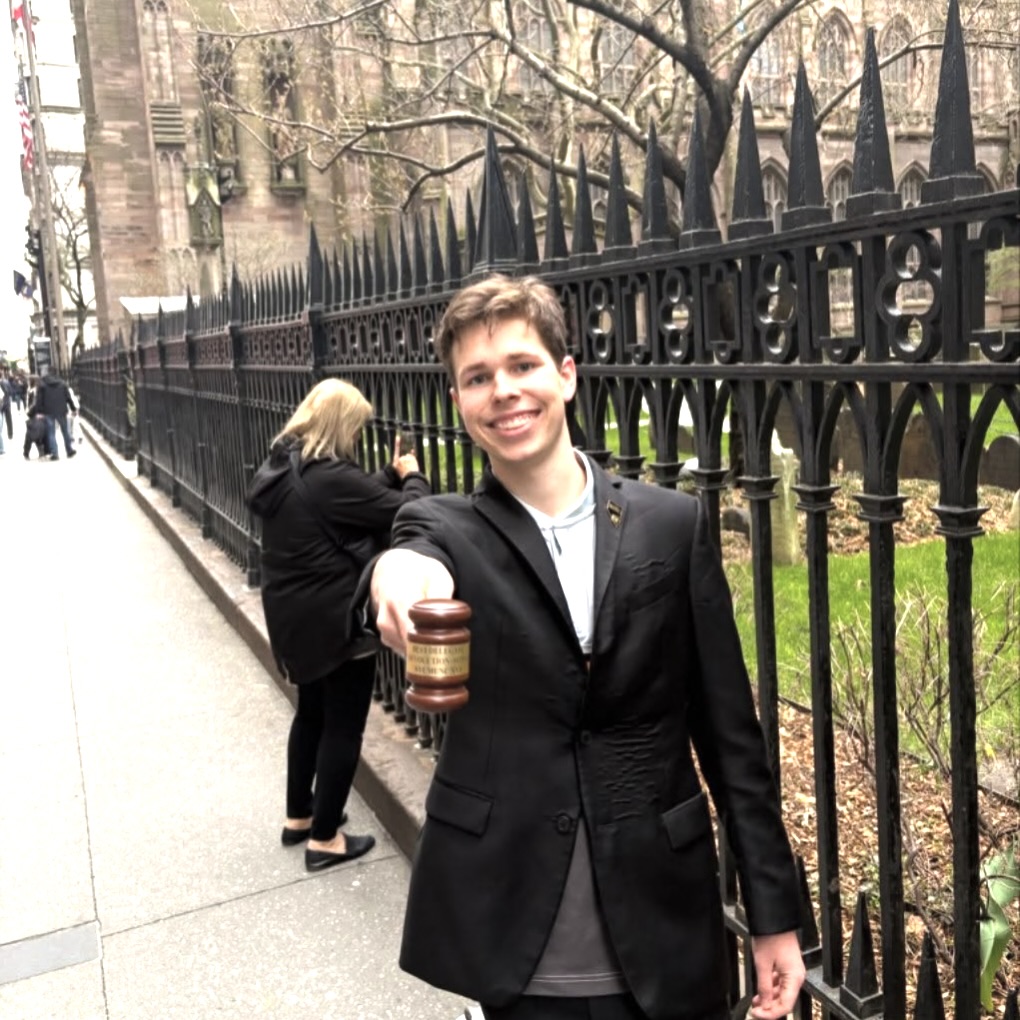Photo: A person in a black suit holds a gavel out toward the camera while standing on a sidewalk next to an ornate black iron fence, with an old stone building and another person in the background.