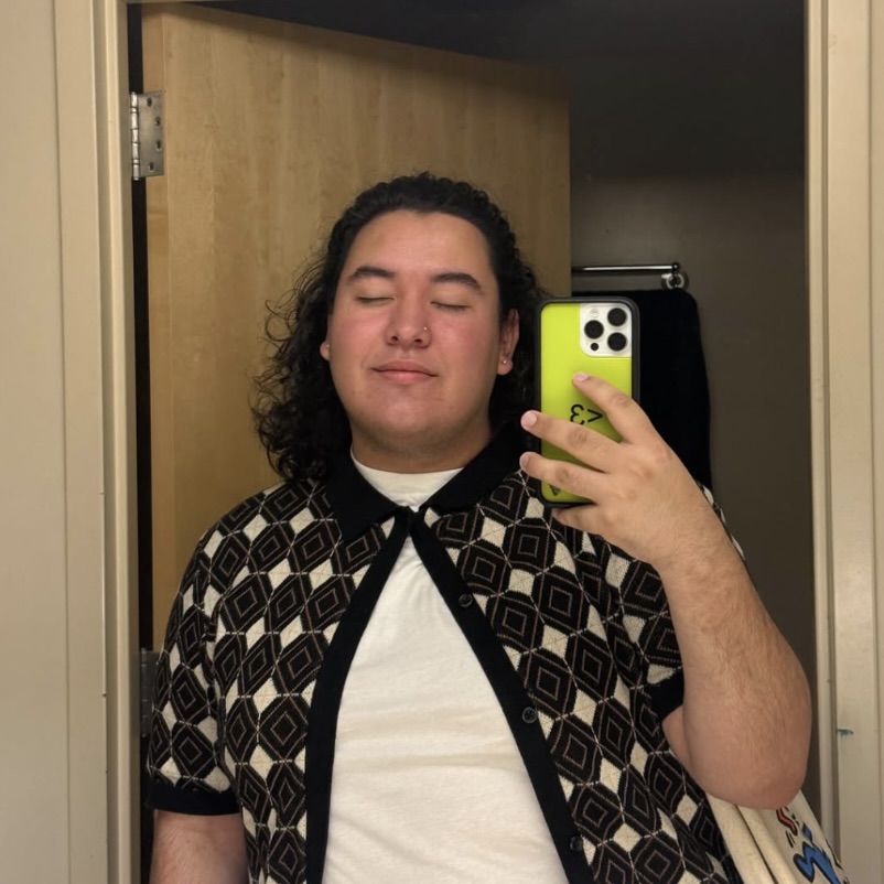 Photo: A person stands in front of a mirror taking a selfie with a neon green phone, wearing a patterned black and white button-up shirt over a white t-shirt. Their eyes are closed.