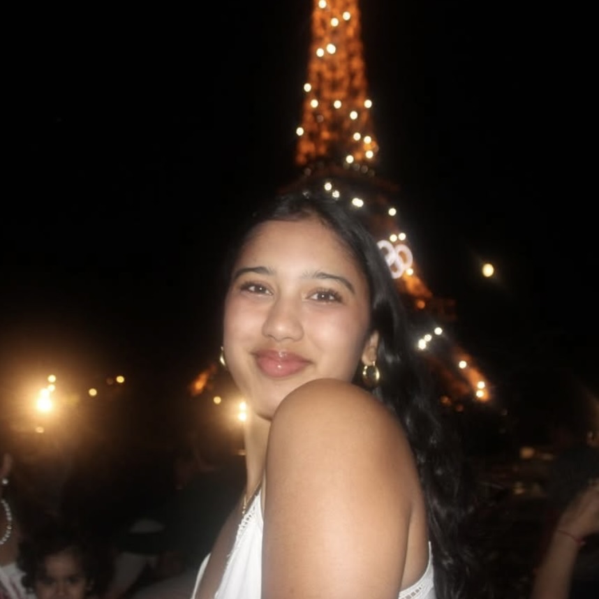 Photo: A person with long, dark wavy hair is standing in front of the illuminated Eiffel Tower at night, wearing a sleeveless white outfit.