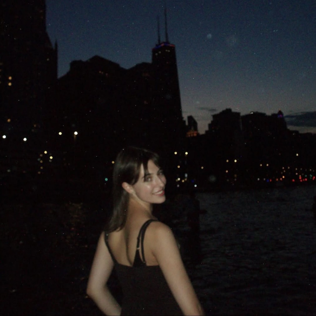 Photo: A person with straight, long hair stands outdoors at night with a city skyline in the background, next to a body of water. The individual is wearing a black sleeveless top.