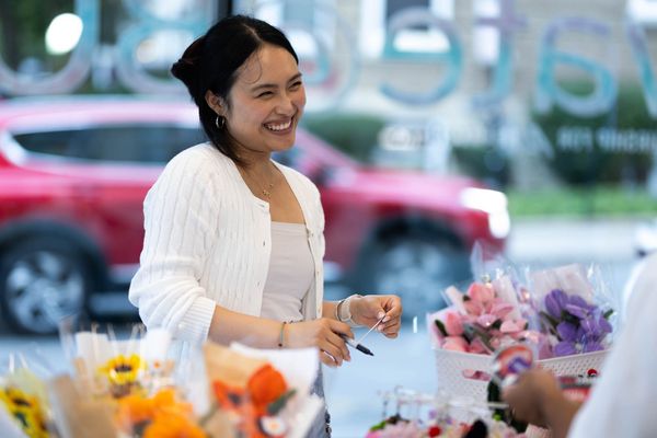 Photo: A woman manning a arts table market smiles as she talks to a customer.
