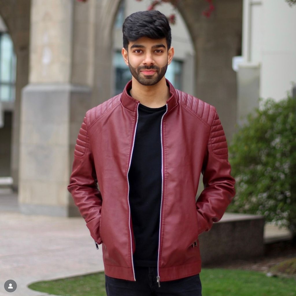 Photo: A person is standing outdoors in front of a stone archway, wearing a maroon zip-up jacket over a black shirt. Their hands are in their jacket pockets.