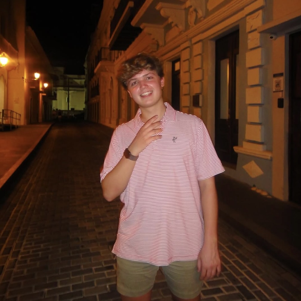 Photo: A person standing on a cobblestone street at night, surrounded by lit buildings painted in light tones. They are wearing a pink and white striped polo shirt, light-colored shorts, and a wristwatch.