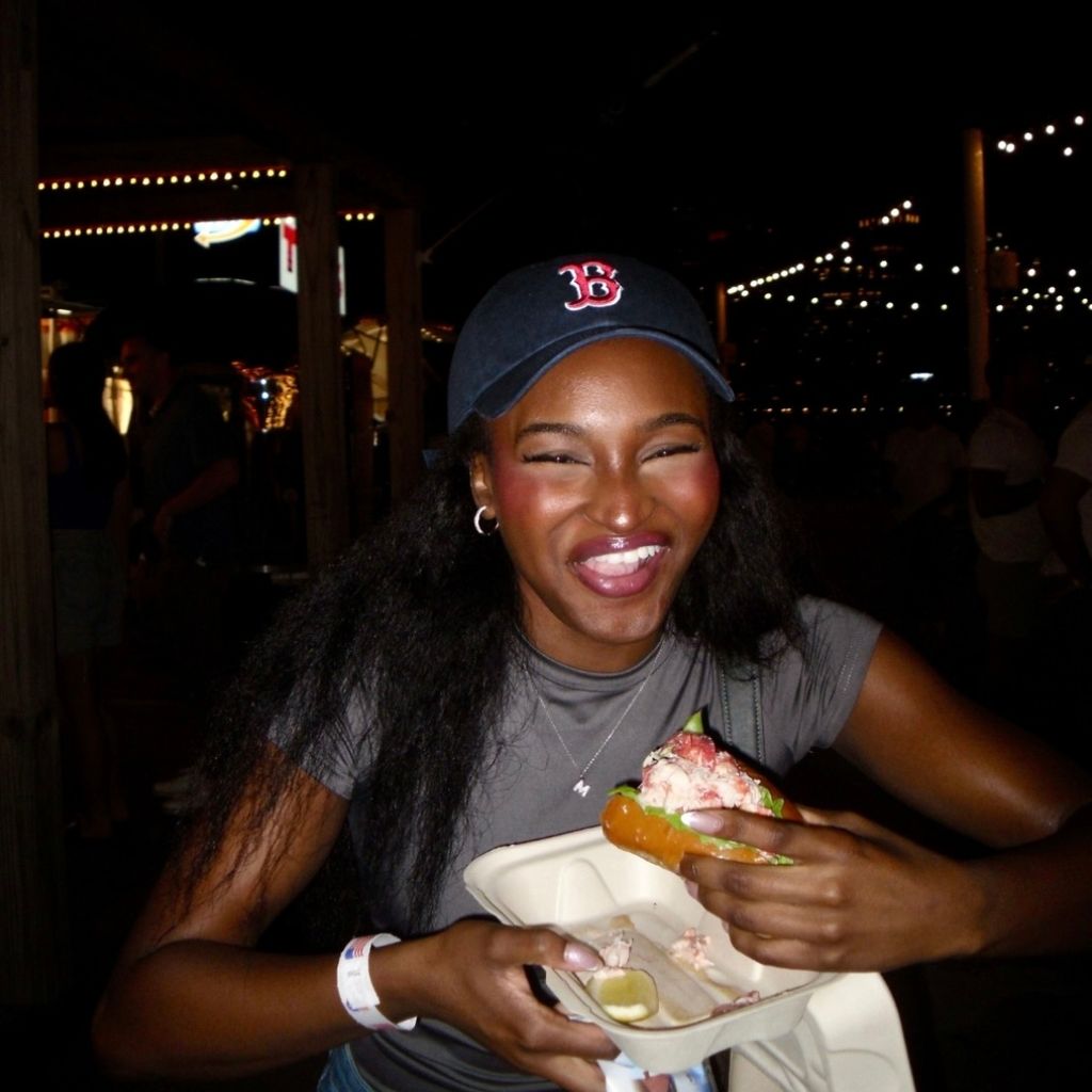 Photo: A person with long hair wearing a navy blue baseball cap with a red "B," hoop earrings, and a gray t-shirt is holding a lobster roll in a takeout container at an outdoor nighttime event with string lights.