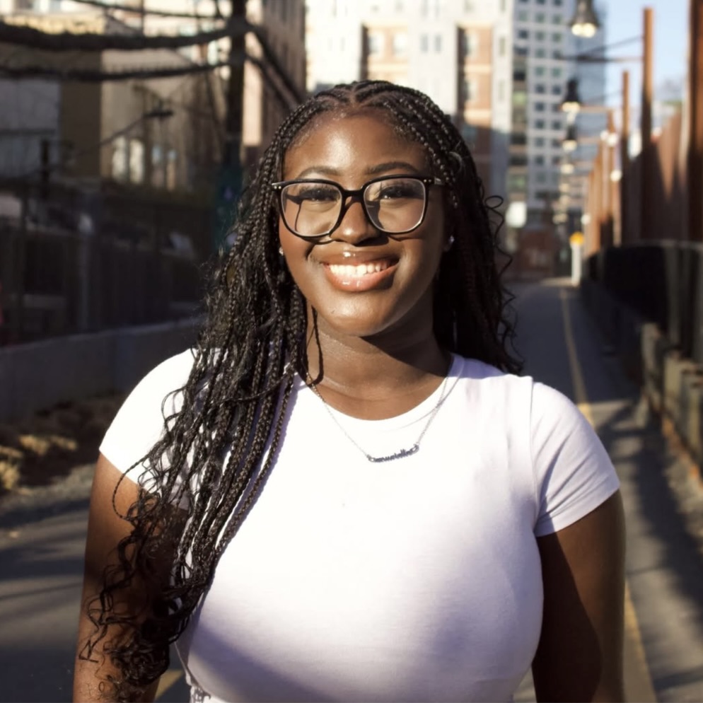 Photo: A person standing on a city street during the day, wearing a white fitted t-shirt and a necklace. Their hair is styled in long braids and the background shows buildings and outdoor lighting.