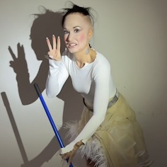 Meme: Person in a white long-sleeve top and beige skirt stands against a wall while holding a broom. Their hand is raised, making a peace sign, with a shadow cast on the wall behind them.
