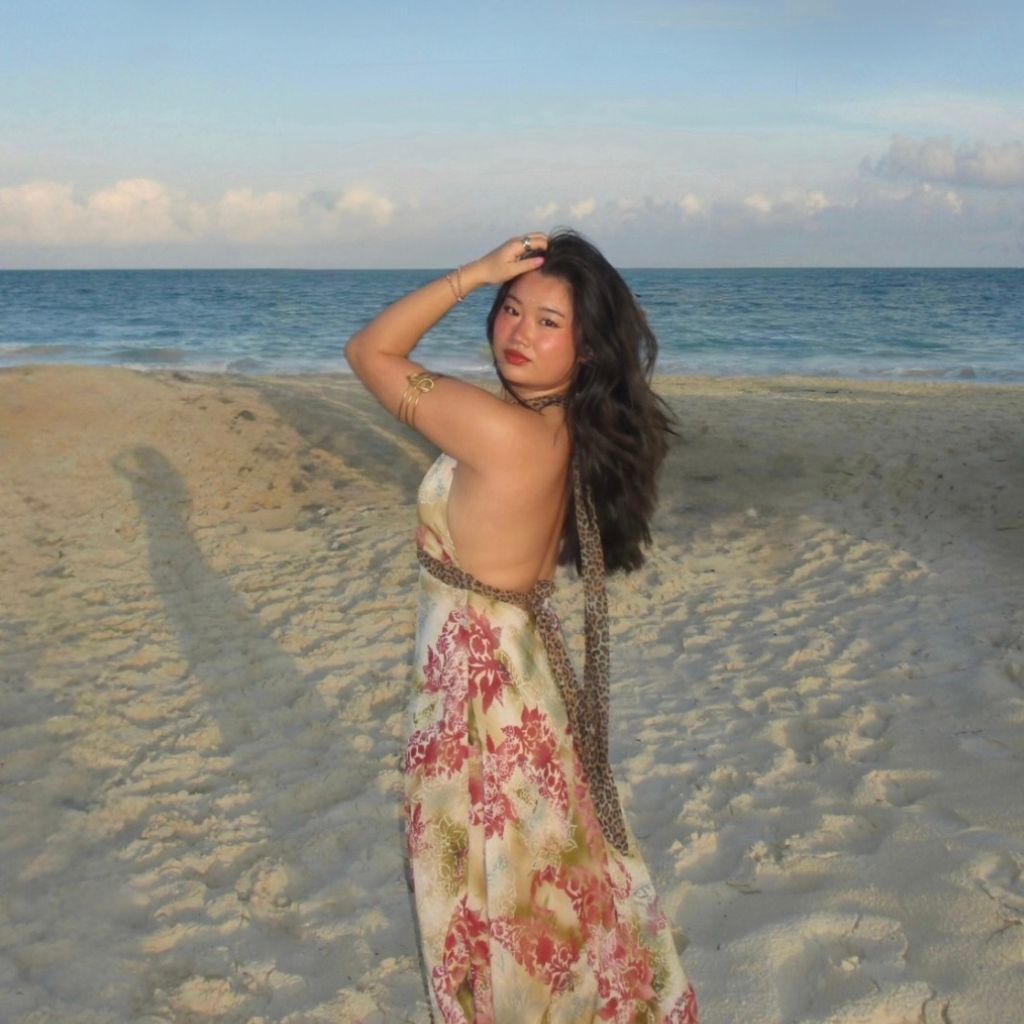 Photo: A person with long, wavy hair stands on a sandy beach near the ocean, wearing a floral maxi dress with gold and brown accessories and holding one hand on their head. The sky is partly cloudy and the water is calm.