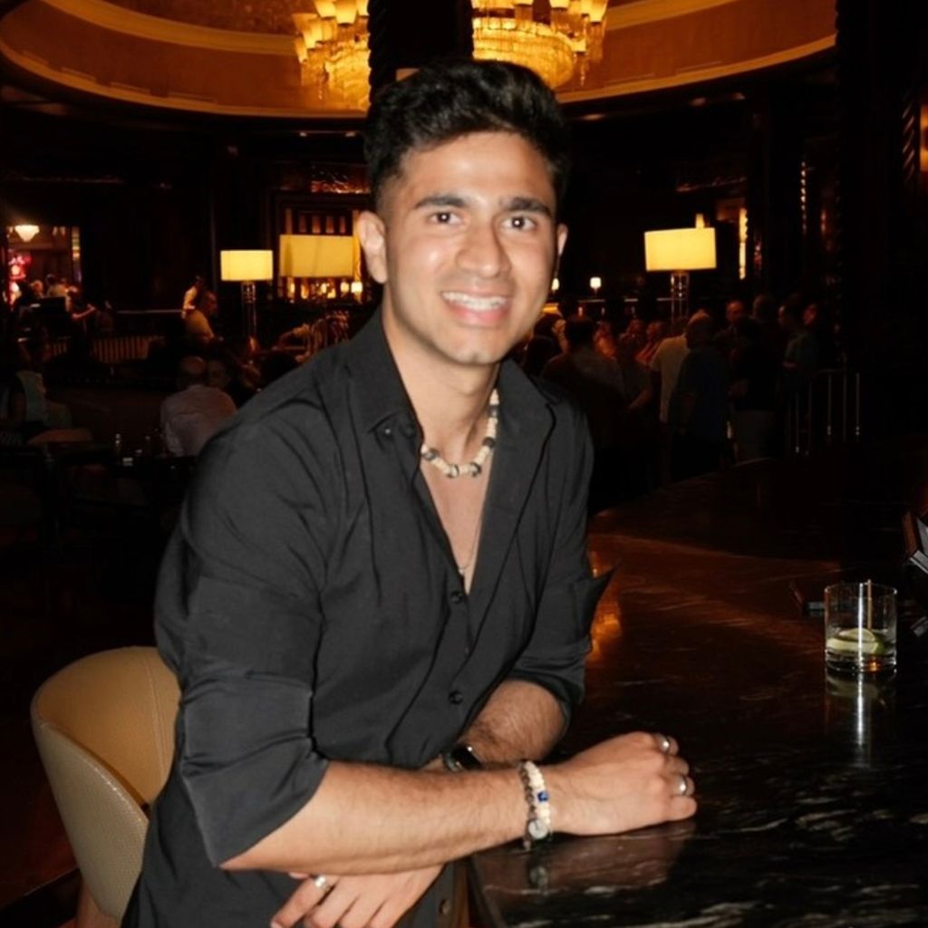Photo: A person is leaning on a dark marble bar counter in a dimly lit, elegant lounge or bar. They are wearing a black button-down shirt with rolled sleeves, a beaded necklace, and bracelets, with a drink on the counter nearby.