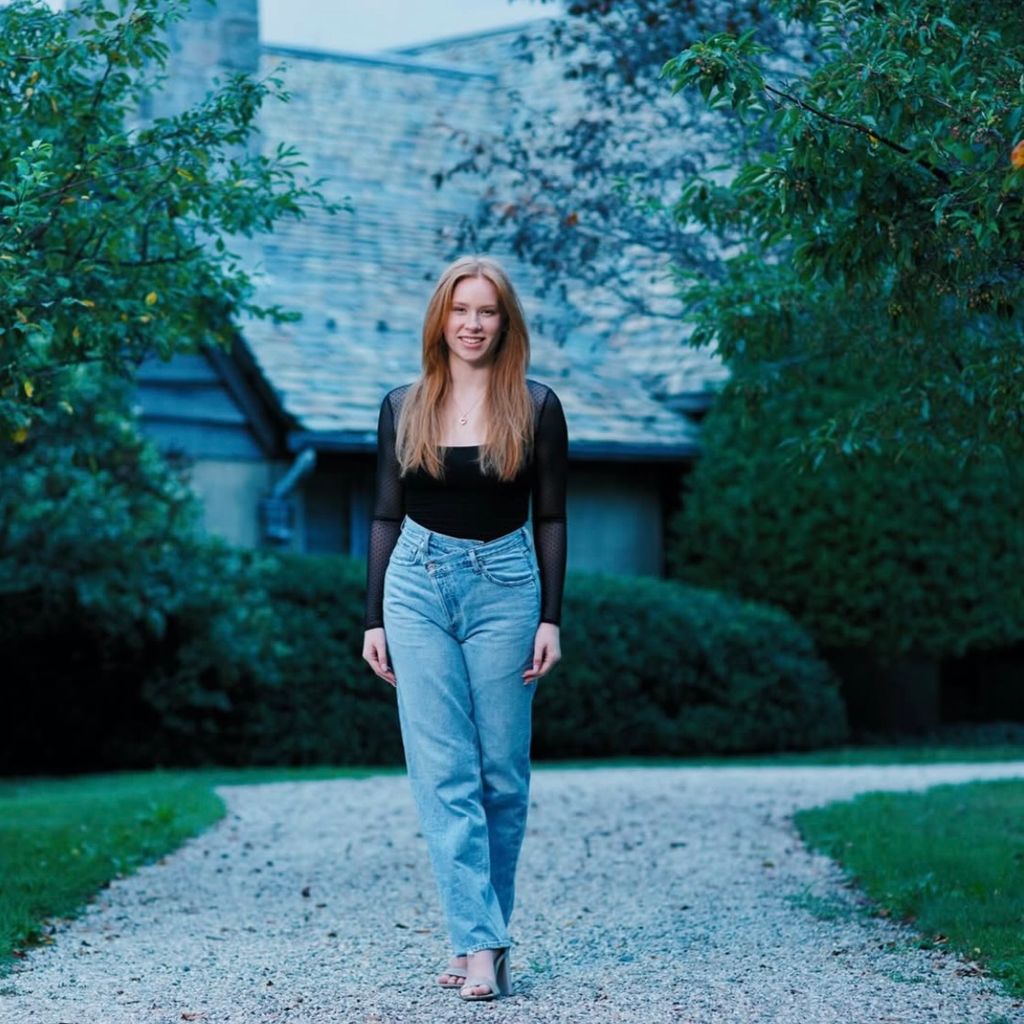 Photo: A person with long, light brown hair is walking in heeled sandals on a gravel path in front of a house surrounded by greenery. They are wearing high-waisted blue jeans and a long-sleeved black top with sheer sleeves.