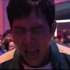 Meme: Individual wearing a dark jacket sits indoors in front of a crowd, with colorful lighting in the background.