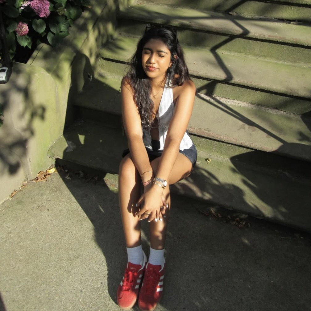 Photo: A person with wavy long hair is sitting in the sunlight on outdoor steps next to a patch of flowers. They are wearing a sleeveless white top, dark shorts, white socks, and bright red Adidas sneakers. Their eyes are closed.