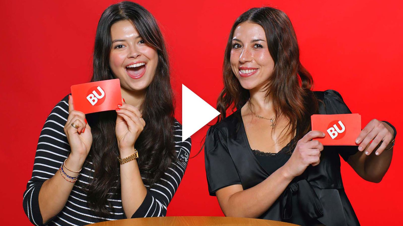 Photo: Two college woman, one wearing stripes on the left and one wearing a black shirt on the right, hold up a red card with BU's logo on the back. They are both smiling wide and are posed in front of a red background. A play button vector is on top of the image.