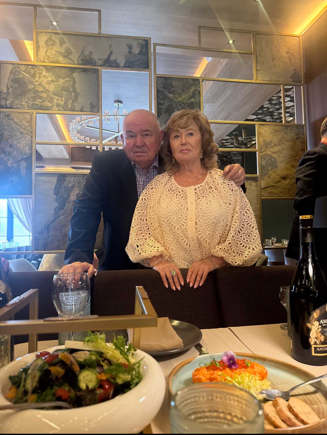 Photo: A well-dressed couple posing together behind a restaurant booth. The man stands slightly behind with one hand on the woman’s shoulder; he wears a dark suit jacket and checked shirt. The woman stands in front wearing a cream-colored lace blouse and a ring on her right hand. A plated salad, a colorful appetizer, glassware, and a wine bottle sit on the table in the foreground. Decorative mirrors and a chandelier are visible on the wall behind them.