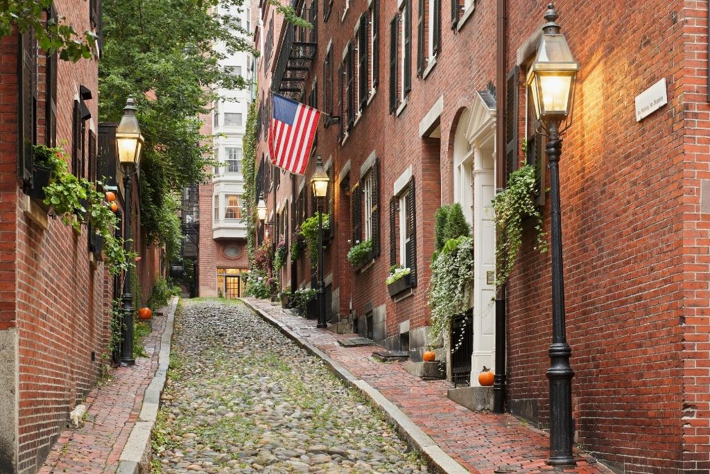 A narrow cobblestone alley in Boston’s Beacon Hill neighborhood lined with red brick buildings, glowing streetlamps, window boxes, pumpkins, and an American flag.