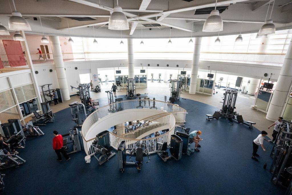 Photo: Boston University's "FitRec" gym, a spacious, multi-level fitness center with weight machines, cardio equipment, a central spiral staircase, and people working out under bright overhead lights.