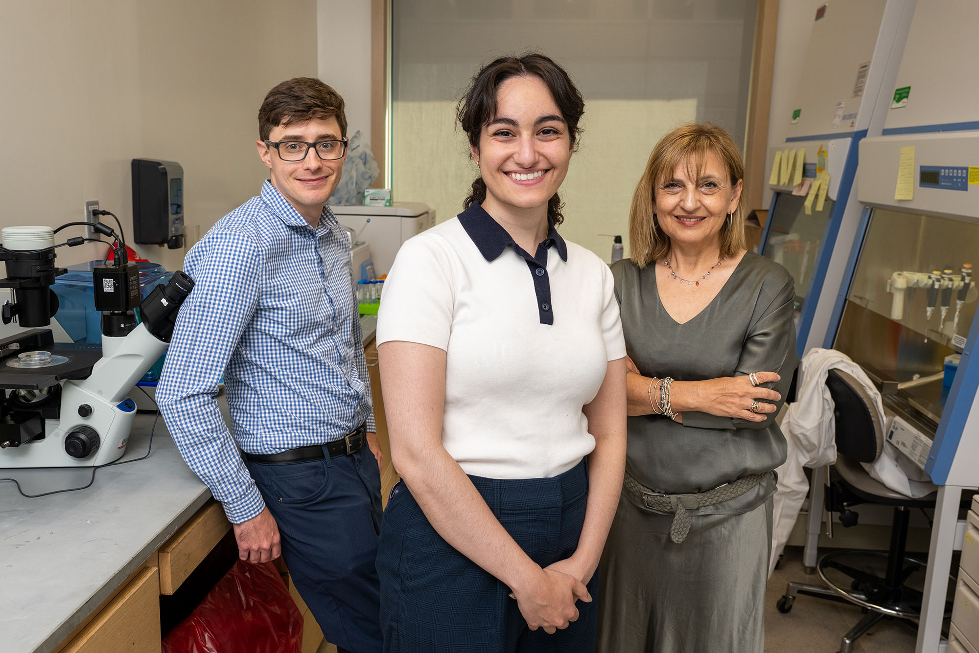 Photo: A group portrait of three smiling adults standing in a laboratory. On the left, a person wearing glasses and a blue-and-white checkered shirt stands beside a large white microscope. In the center, a person with dark hair wears a white shirt with a dark navy collar and cuffs. On the right, a person with shoulder-length light brown hair wears a gray blouse and jewelry. Laboratory equipment, countertops, containers, notes, and a glass-fronted instrument cabinet fill the background.