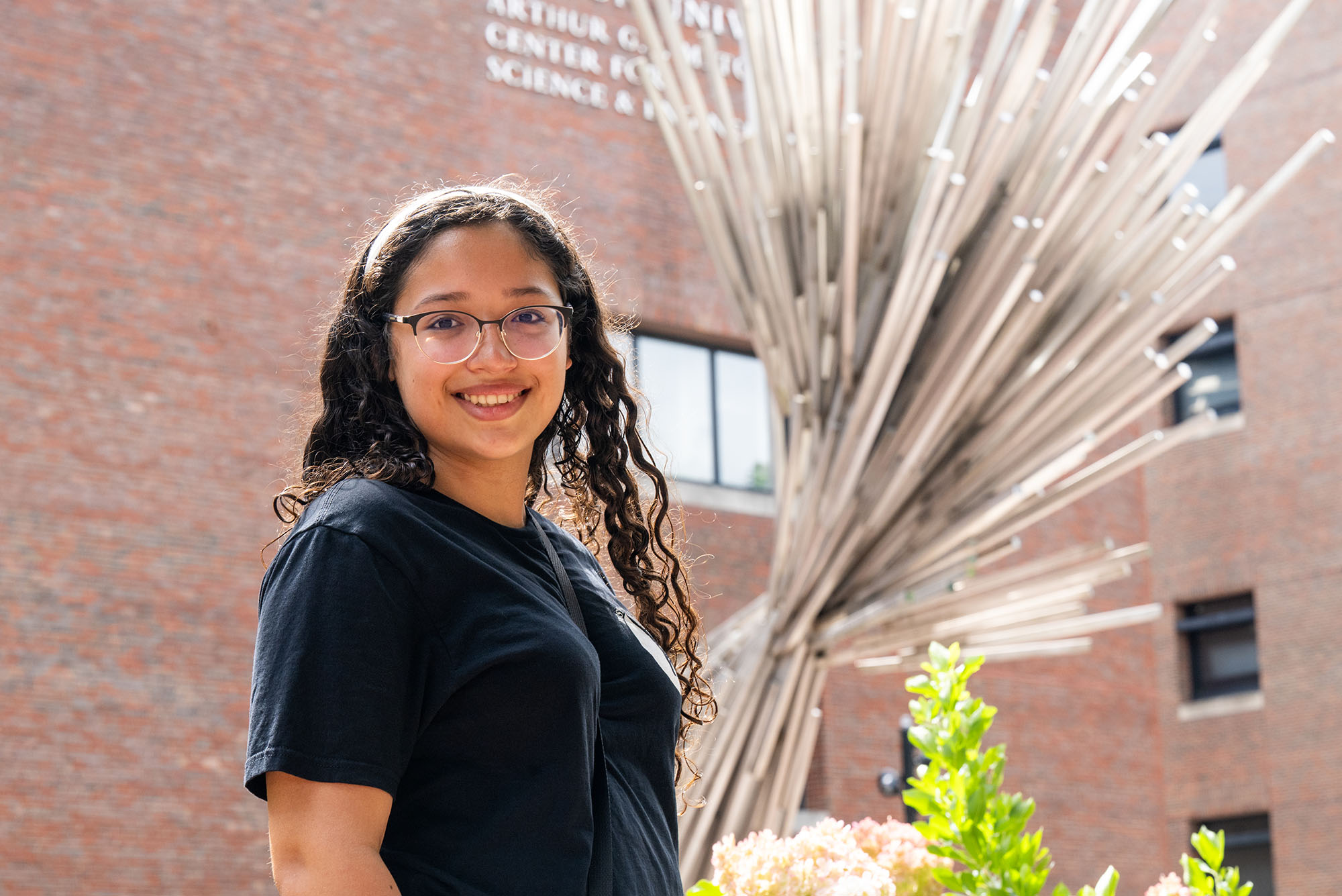 Photo: Smiling college student in glasses standing beside a large radiating metal-rod sculpture in a brick courtyard.