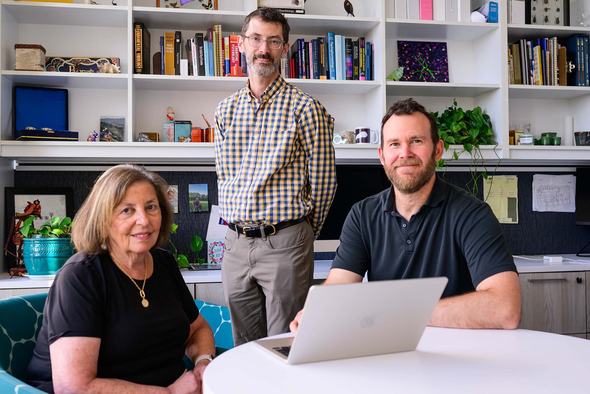 Photo: Three adults pose in a room with bookshelves, plants, and an open laptop on a white table.