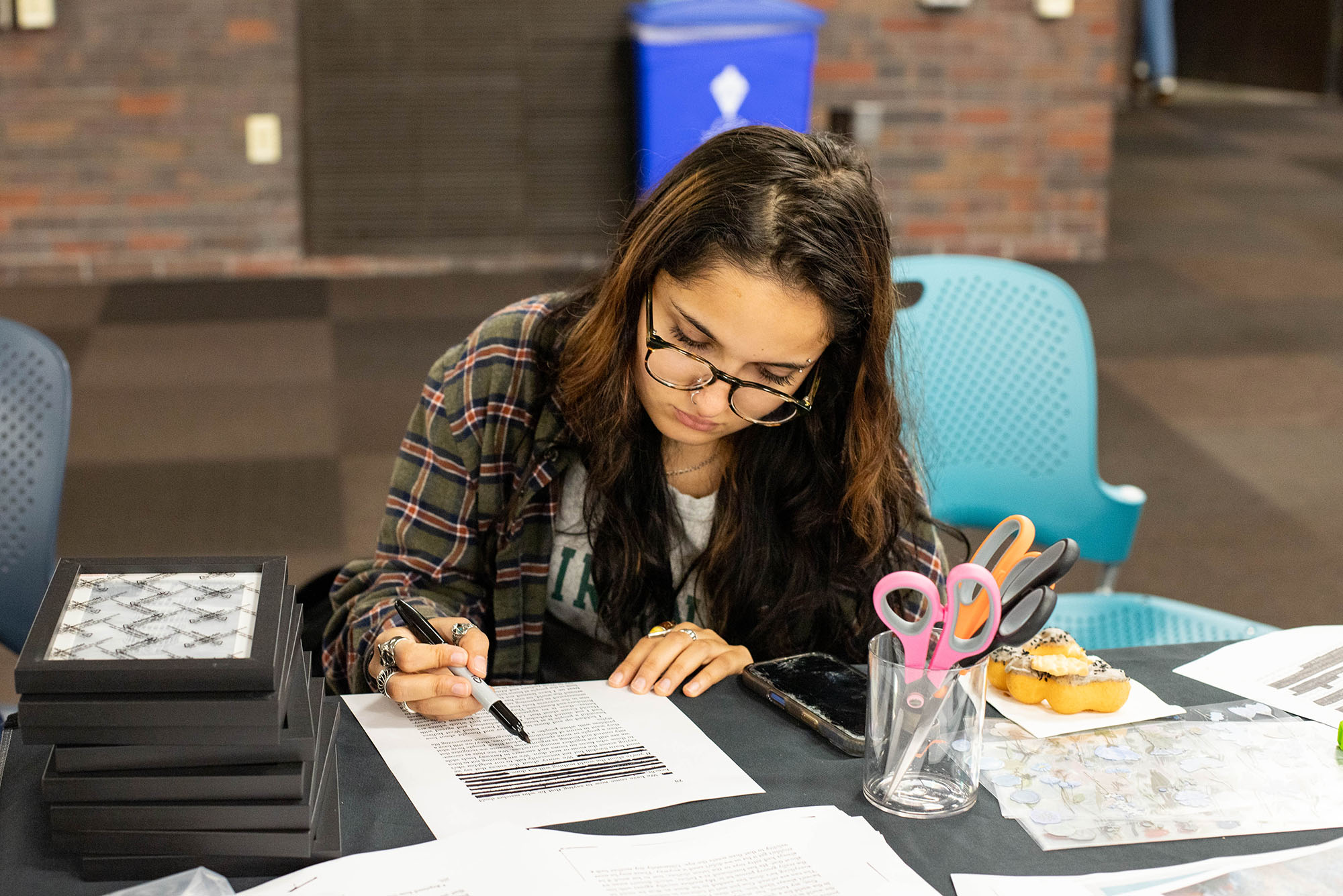 Photo: Person wearing glasses writes on a document at a table surrounded by papers, scissors, a phone, and pastries.