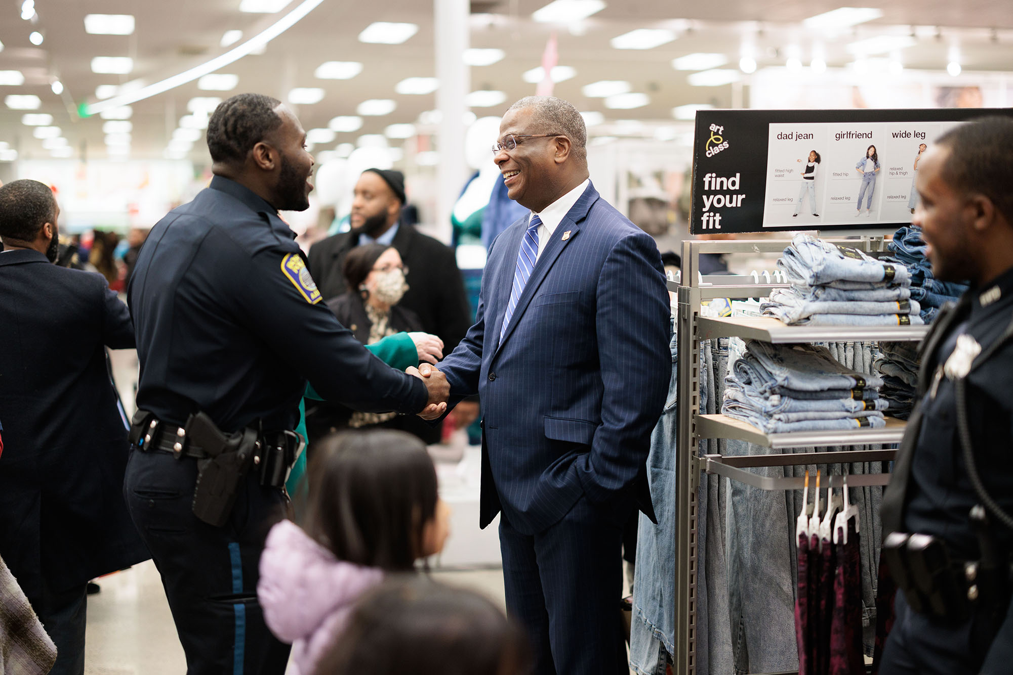 Photo: Al Colbourne, B2 Officer, left, and Xavier Hill, C6 Officer, right, speak to newly appointed police commissioner Michael Cox, middle, at the 11th annual "Shop with a Cop" event at Target in the South Bay shopping plaza in Dorchester on December 13th. One Black man in a Boston police officer uniform shakes hands with another Black man wearing glasses, a navy blue suit, and lighter blue striped tie. They both smile as various other people of color look on. Another Black officer in uniform looks on from the right.