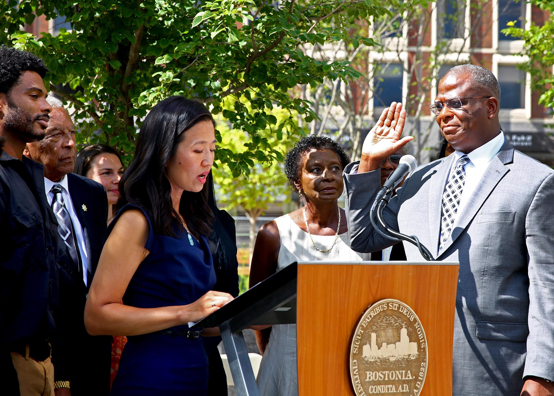 Photo: Boston, MA - August 15: With his family gathered around him, Michael A. Cox is sworn in as Bostons new Police Commissioner by Mayor Michelle Wu. An Asian woman wearing a navy blue summer dress reads off a podium as a Black man to her right, wearing a grey suit and striped dark grey tie, raises his right hand as he listens on. Various other Black people stand next to and behind the Asian woman as they watch on.