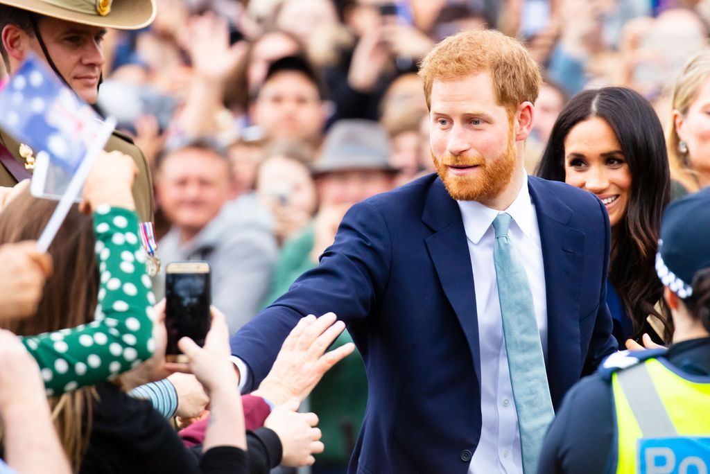 Photo: Prince Harry and Megan are amongst a large crowd, shaking hands and exchanging pleasantries. Harry wears a blue suit with a blue tie and Meghan's outfit is not visible, but her hair is down and she is smiling.