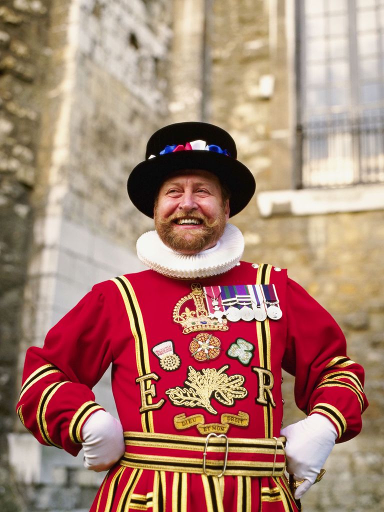 Photo: A kings guard has his arms on his hips while he looks to the left. He is smiling in his traditional liveries which are red with white and gold detailing. He also wears a black hat.
