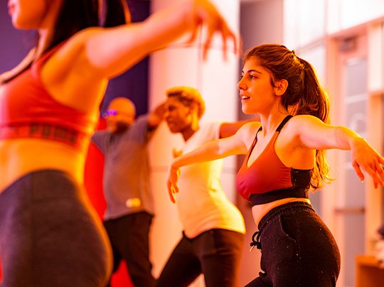 People participating in a group fitness class, moving in unison with arms raised, in a brightly lit studio.
