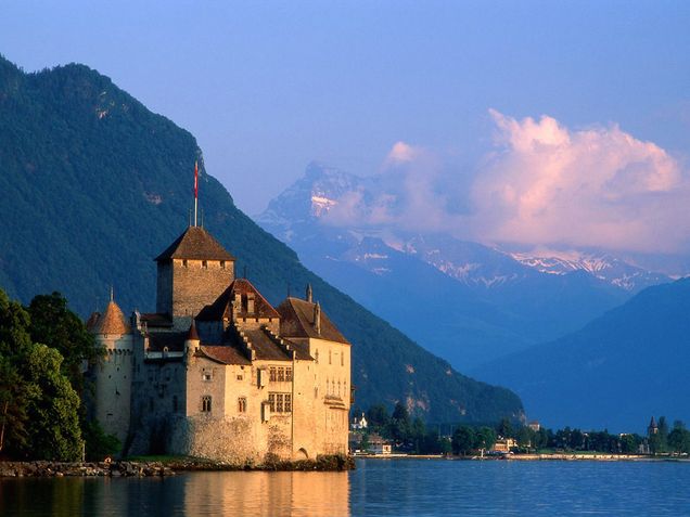 The image shows the lakeside Chateau de Chillon, a historical site near Montreux, Switzerland. There are clouds and mountains in the background of the photo, and the sky is dark blue, turning to dusk.
