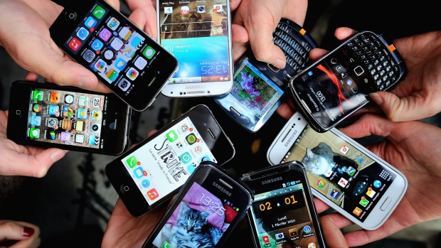 Nine different types of mobile phones showing various homescreens being held out by different people's hands.
