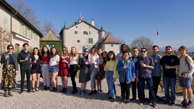A group of 19 BU students are gathered during a wine-tasting event at Chateau du Crest.  The Chateau is located in the countryside near Geneva, and is visible behind the students.  The sky is blue and cloudless in the background.