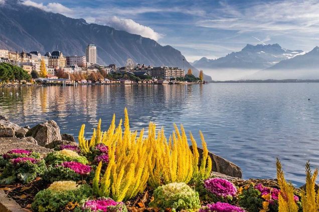 The image is a cityscape of Montreux, Switzerland. In the foreground, there are brightly-colored flowers and landscaping along a lakeside promenade. Montreux is in the middle ground, with mountains, clouds and blue sky in the background.
