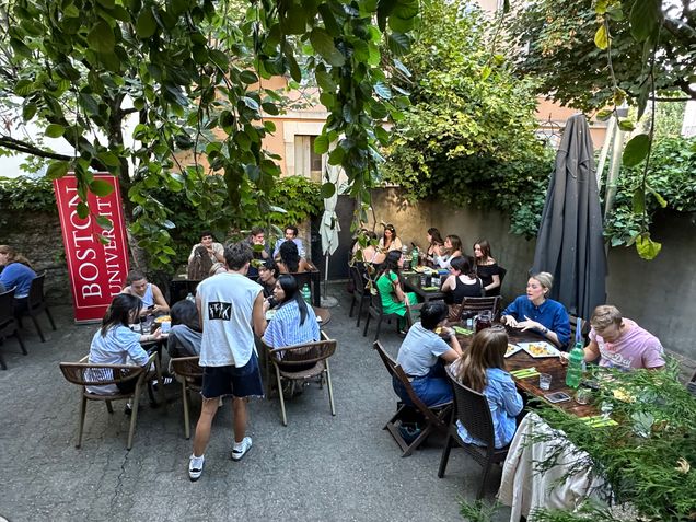 A group of 35 BU students and staff at the Fall 2024 Welcome Dinner. The setting is a leafy courtyard, with a large red BU banner and people distributed between five tables.