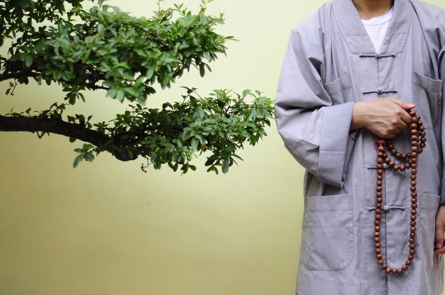 2016 GP Photo Contest Finalist monk with preyer beads standing against a wall next to a tree