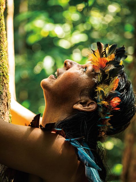Woman with feathered wreath in her hair hugging and looking up at a tree in the Amazon Rain Forrest. 2026 GP Photo Contest finalist