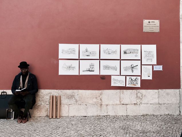 Artist sitting against a wall reading a book near a number of his displayed sketches on the wall. 2026 GP Photo Contest finalist.