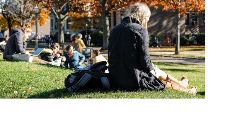 A student sits in the grass and looks at her phone with the colorful fall leaves behind her.