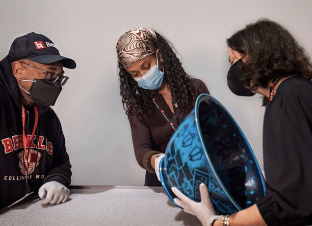 Three people stand, masked looking on the bottom of a large blue bowl.