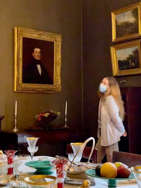 Erin Sutton-Beals, wearing a face mask, poses with a portrait painting of a man on the wall of an historic home museum.
