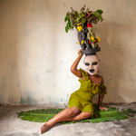 a figure in a green dress wears a very large white mask with figures of people atop their head. the figure leans against a wall, holding up the weight of the mask while sitting sideways on the floor.