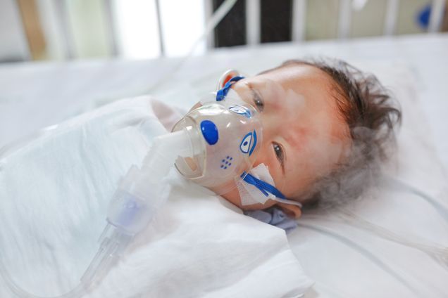 Toddler boy using nebulizer to cure asthma or pneumonia disease . Sick baby boy rest on patients bed and has inhalation therapy by the mask of inhaler. Respiratory Syncytial Virus (RSV).