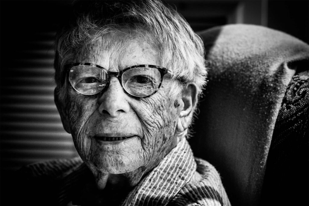 Photo: an up-close portrait of a centenarian wearing glasses and a sweater. Photo is black and white