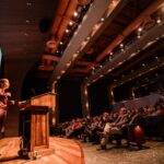 Photo: A woman in a suit stands at a podium in front of a crowded auditorium, giving a lecture