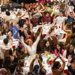 Photo: A picture of many people celebrating with their arms raised as they sit around round tables