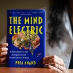 Photo: Pria Anand holding up her book "The Mind Electric," which features a vibrant blue cover and an illustration of the brain with flowers making up different parts