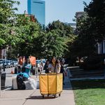 Photo: A BU freshman rolling a large, yellow bin during move-in with the Boston skyline behind her