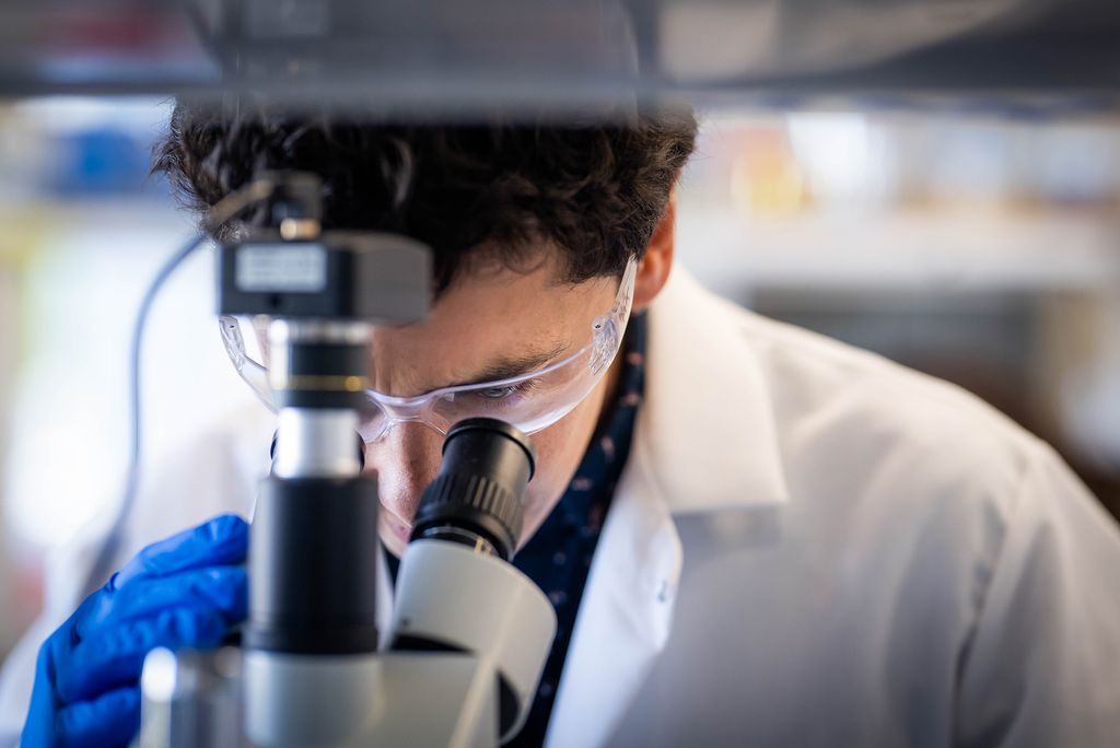 Photo: Miguel Jimenez, an engineer at BU, looking into a microscope while wearing a lab coat and safety glasses.